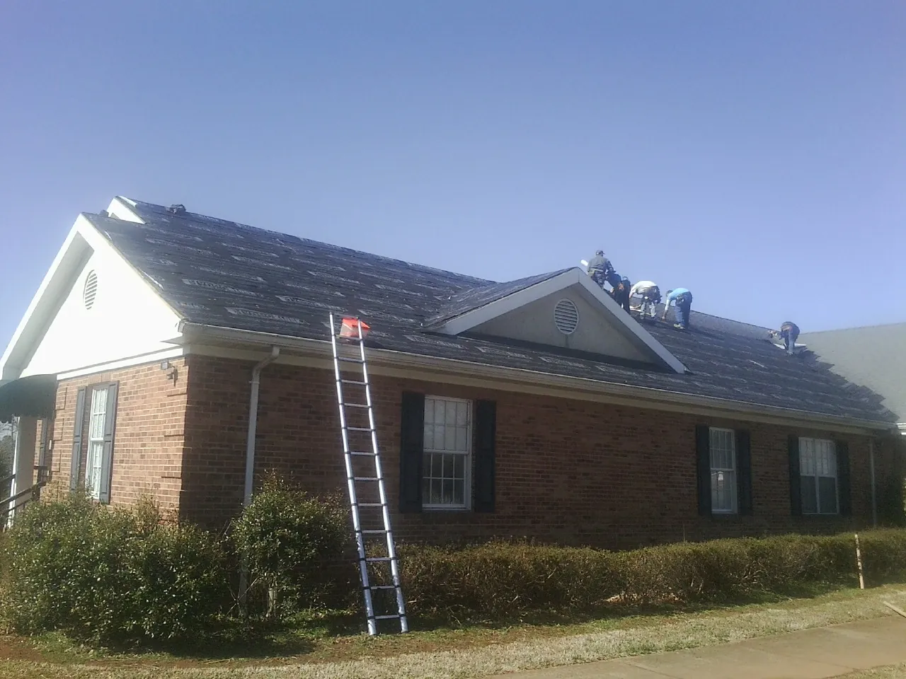 Crew tearing shingles off a brick ranch roof with a ladder set — Emergency Roof Repair work by our crew in Center Junction