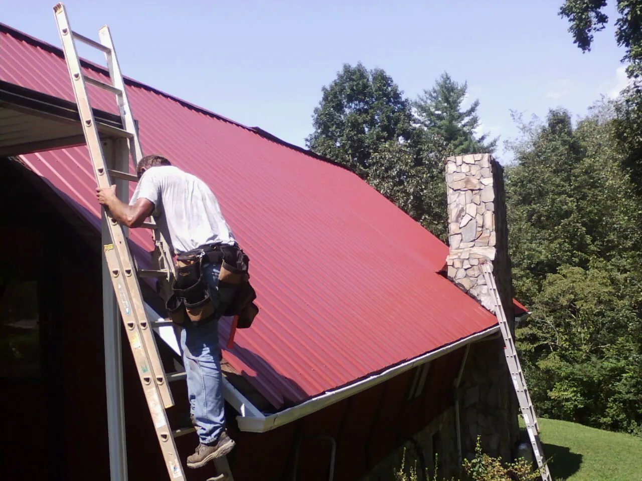 Roofer on a ladder beside a red metal roof and stone chimney — Soffit & Fascia Repair by Roofing Brothers in Center Junction